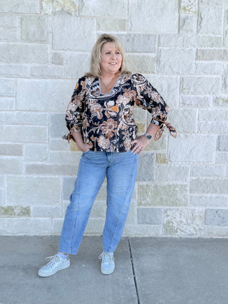 Woman wearing a floral blouse and blue jeans standing against a stone wall.