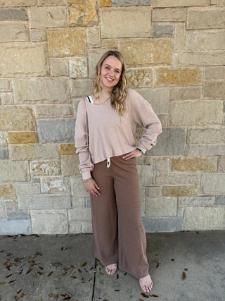 Woman wearing a beige sweater and brown pants standing against a stone wall.