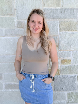 Woman wearing a beige tank top and blue denim skirt standing against a stone wall.