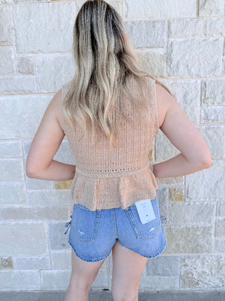 Person wearing a beige knit top and denim shorts with a visible brand tag, standing against a stone wall.
