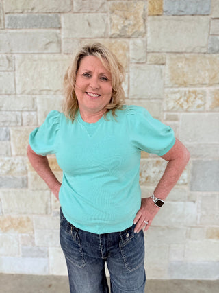 Woman wearing a light blue top and jeans standing against a stone wall.