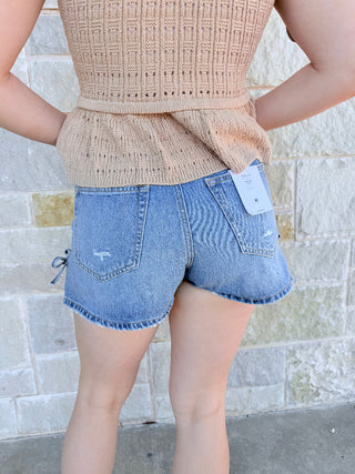Person wearing a beige knit top and blue denim shorts with a stone wall background