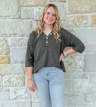 Woman wearing a dark gray shirt and light blue jeans standing against a stone wall.