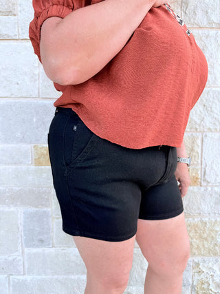 Person wearing a rust-colored top and black shorts against a tiled wall.