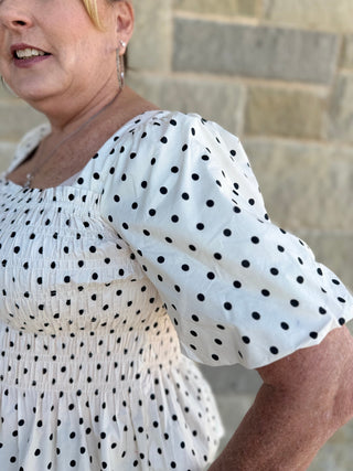 Woman wearing a white blouse with black polka dots against a stone wall.