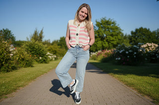 Person wearing a pink and white striped vest, light blue jeans, and black sneakers against a stone wall.