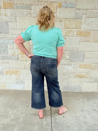 Person wearing a light green t-shirt and blue jeans standing against a stone wall.