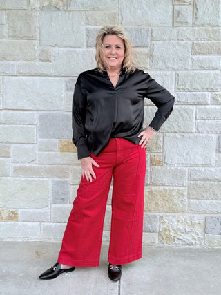 Woman wearing a black blouse and red pants standing against a stone wall.