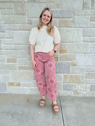 Woman wearing a white top and pink patterned pants standing against a stone wall.