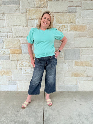 Woman wearing a light blue shirt and dark jeans standing against a stone wall.