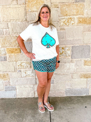 Woman wearing a white t-shirt with a blue graphic design and patterned shorts standing against a stone wall.