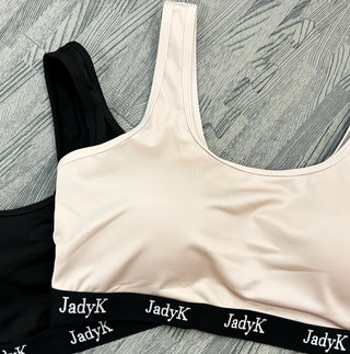 Beige sports bra with black band featuring 'JadyK' branding on a textured gray surface.
