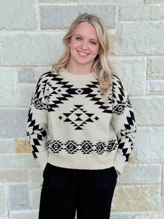 Woman wearing a cream sweater with black geometric patterns against a stone wall.