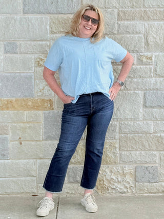 Person wearing a light blue shirt and dark jeans standing against a stone wall.