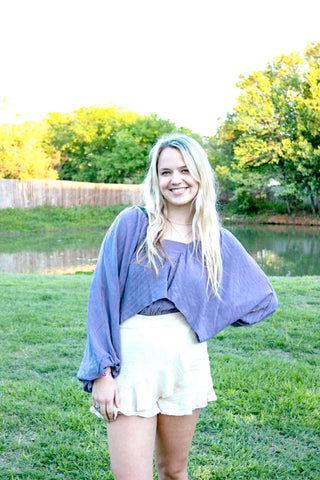 Woman wearing a purple sweater over a white top and skirt standing in a grassy area with trees and water in the background.