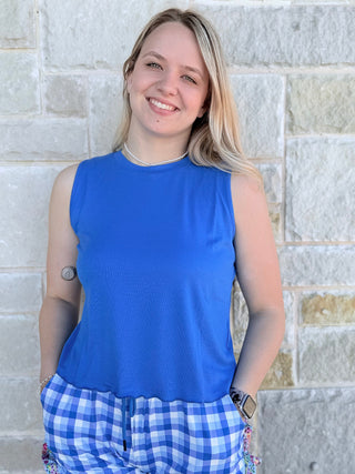 Person wearing a blue sleeveless top and plaid shorts standing against a stone wall.