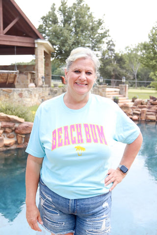 A woman standing by a pool, wearing a blue t-shirt with the text 'BEACH BUM' printed on the front.
