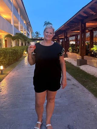 A woman standing on a pathway at dusk, wearing a black boxy cut top with pearl sleeves and a front tie short, holding a glass of orange juice.
