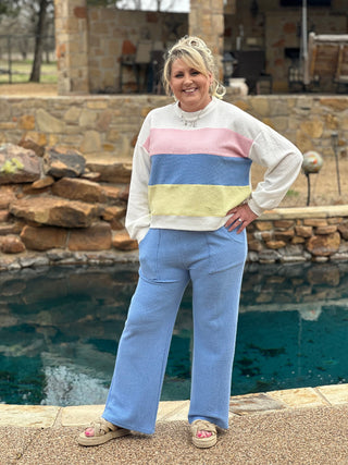 A person wearing a color block long sleeve top with round neck, standing near a pool. The top has horizontal stripes in pink, yellow, and blue colors.