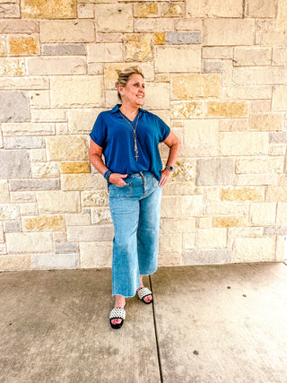 A woman standing with one hand against a stone wall, wearing a blue top and blue jeans with a front pocket, with her foot raised on a stone surface.