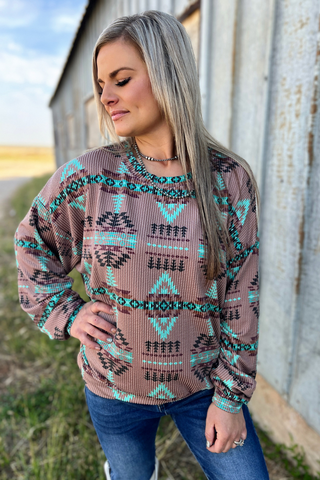 A woman standing outdoors wearing a brown and multicolored geometric patterned sweater and blue jeans.