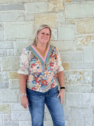 Woman wearing a colorful floral blouse and jeans standing against a stone wall.
