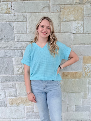 Woman wearing a light blue textured shirt and jeans standing against a stone wall.