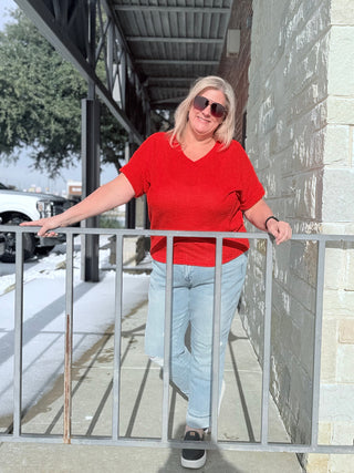 Woman in a red shirt and sunglasses standing on a sidewalk next to a building.