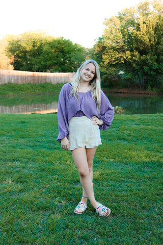 Woman in a purple sweater and white skirt standing on grass with trees in the background