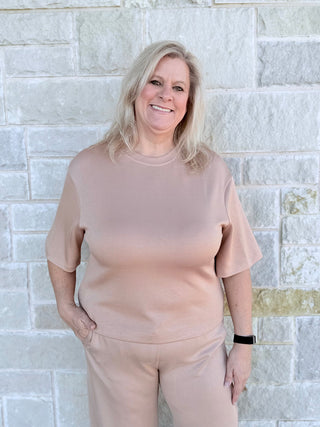 Woman wearing a beige outfit standing against a light stone wall.