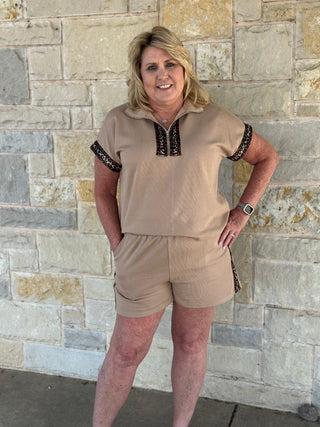 Woman wearing a beige outfit with black trim against a stone wall.