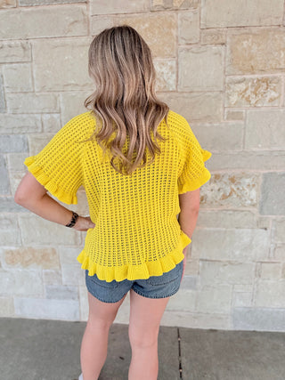 Person wearing a yellow crochet top and denim shorts standing against a stone wall.