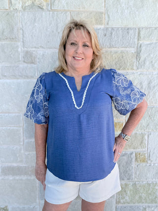 Woman wearing a blue top with white lace sleeves against a stone wall.