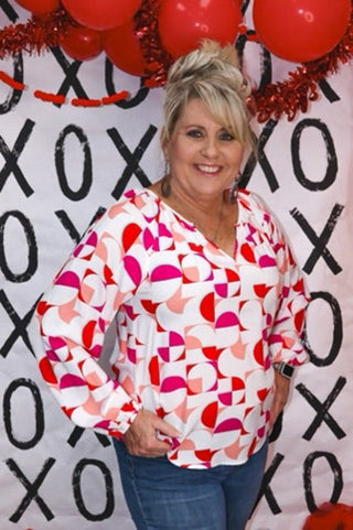 Woman wearing a colorful geometric-patterned shirt in front of a decorative background with 'XOXO' text.