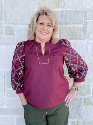 Woman wearing a burgundy blouse with patterned sleeves against a stone wall.