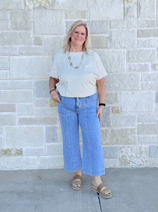 Woman wearing a white top and blue jeans standing against a stone wall.