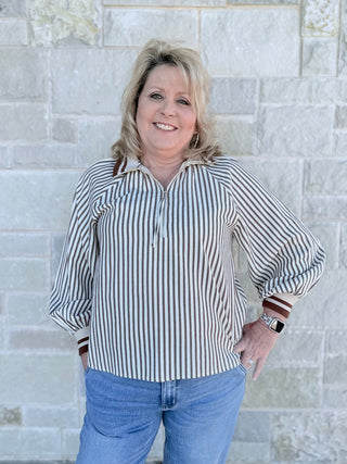 Woman wearing a striped shirt and jeans standing against a light stone wall.