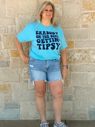Woman wearing a blue t-shirt with text and denim shorts standing against a stone wall.