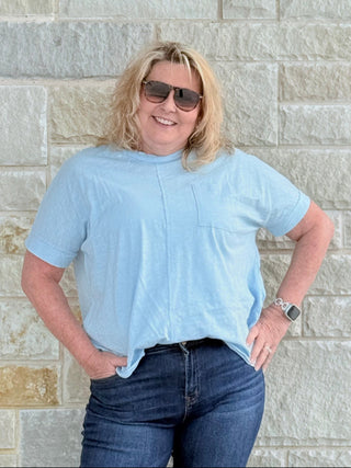 Person wearing a light blue shirt and sunglasses standing against a stone wall.