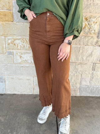 Person wearing brown pants and white sneakers standing against a stone wall.