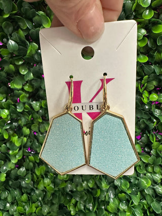 A pair of turquoise geometric earrings dangle on a white card, set against a green leafy background.