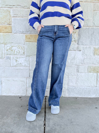 Person wearing a blue and white striped sweater and blue jeans standing against a stone wall.