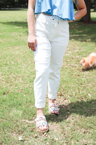 Person wearing white pants and colorful sandals standing on grass with a dog in the background