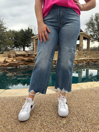 A person wearing high-waisted, rigid denim jeans with a wide leg and a crop cut above the ankle, paired with a pink top and white sneakers.