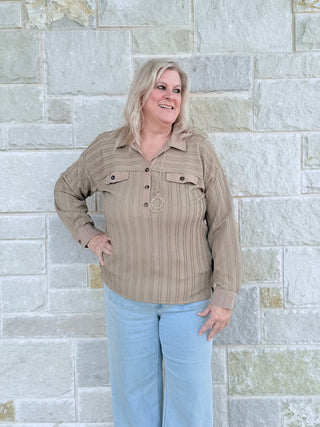 Woman wearing a beige striped shirt and light blue jeans standing against a stone wall.