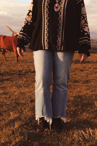 Person wearing a black embroidered top and light blue jeans standing in a field with a cow in the background.