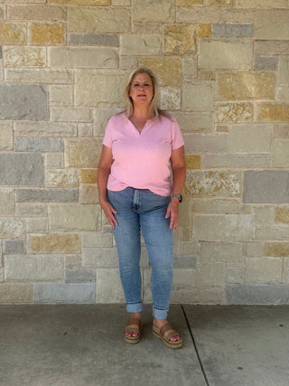 Woman wearing a pink shirt and blue jeans standing against a stone wall.