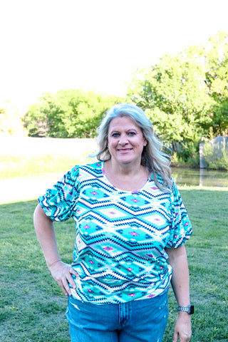 Woman wearing a colorful patterned shirt standing outdoors with trees and water in the background