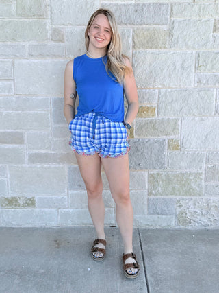 Woman wearing a blue sleeveless top and checkered shorts standing against a stone wall.