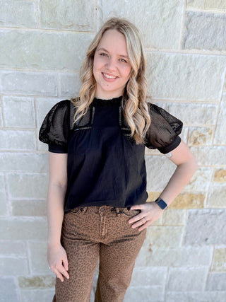 Woman wearing a black top with sheer sleeves against a stone wall.